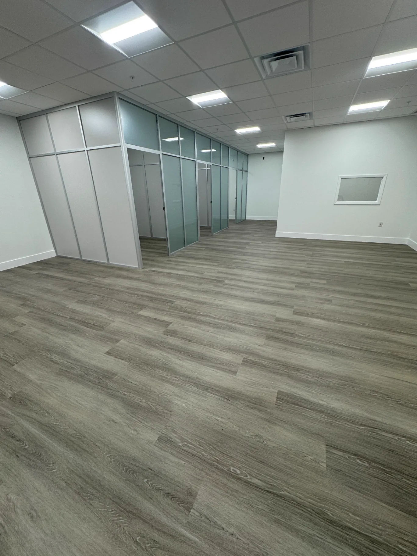 Large open commercial area in Miami with warm-gray wood-look flooring — glass-partitioned private offices in the background.
