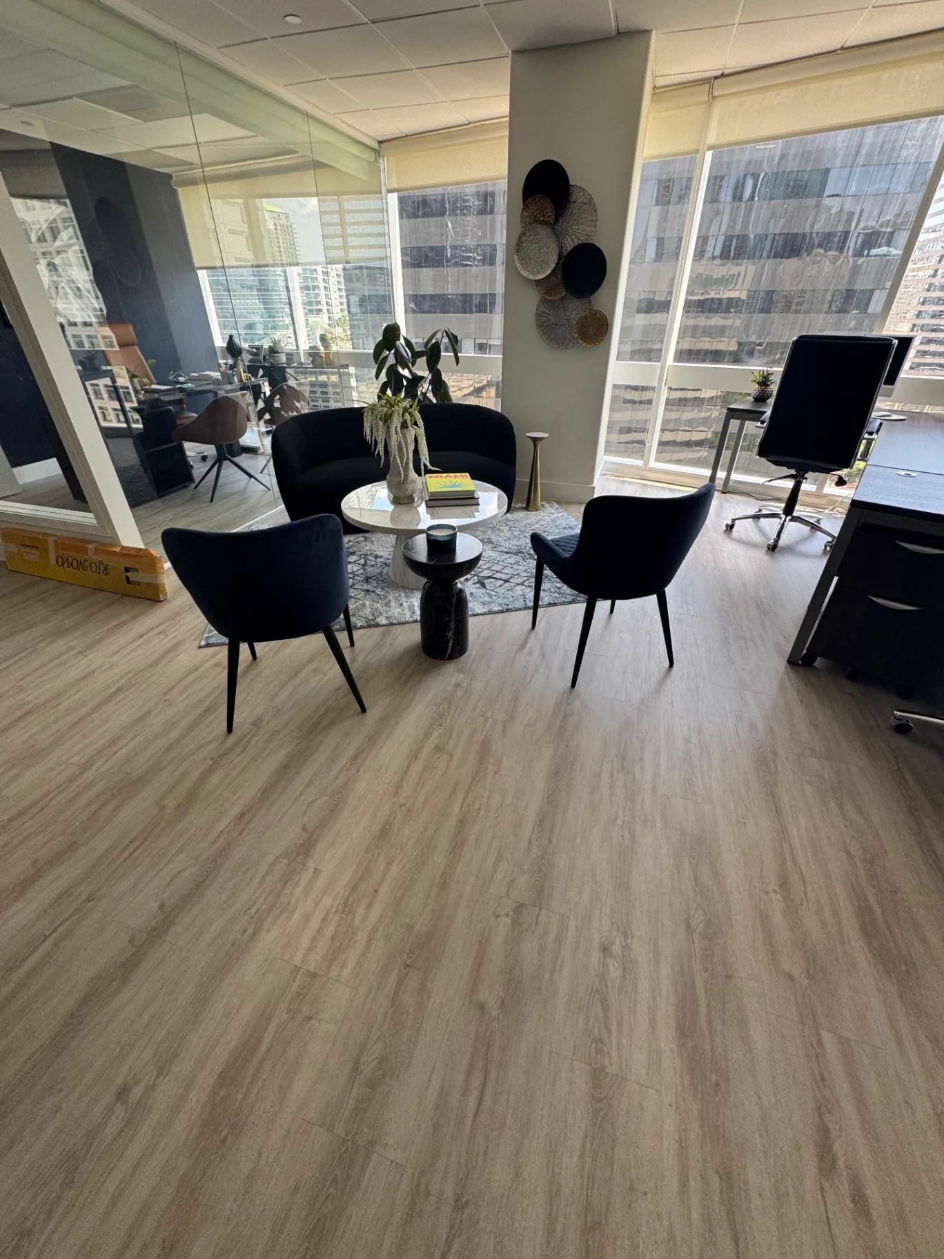 Miami office lounge corner with warm-gray commercial wood-look flooring installed by Colors Wood Floor Corp; floor-to-ceiling windows in the background.