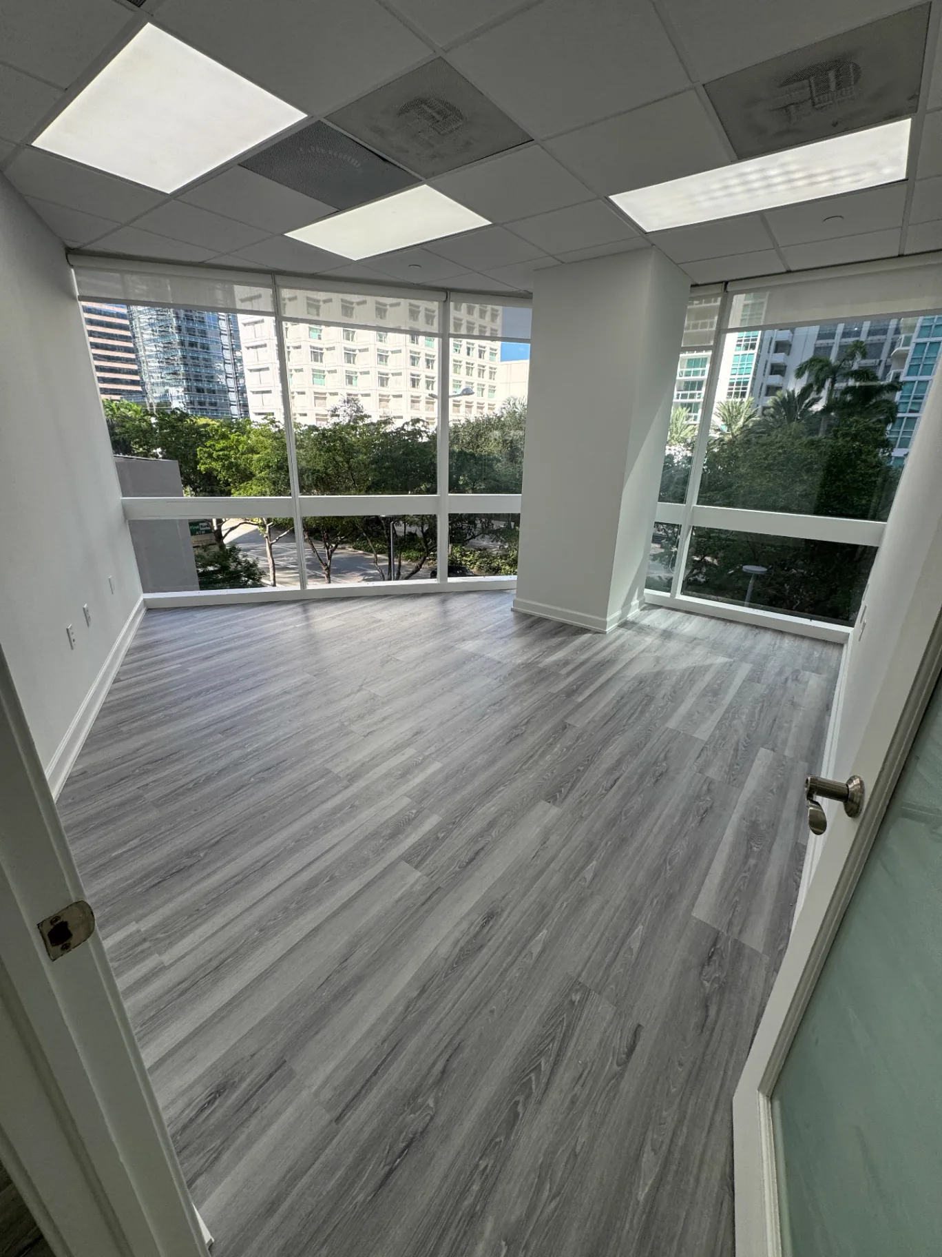 Corner office with full-height Miami city view and light gray wood-look flooring installed by Colors Wood Floor Corp.