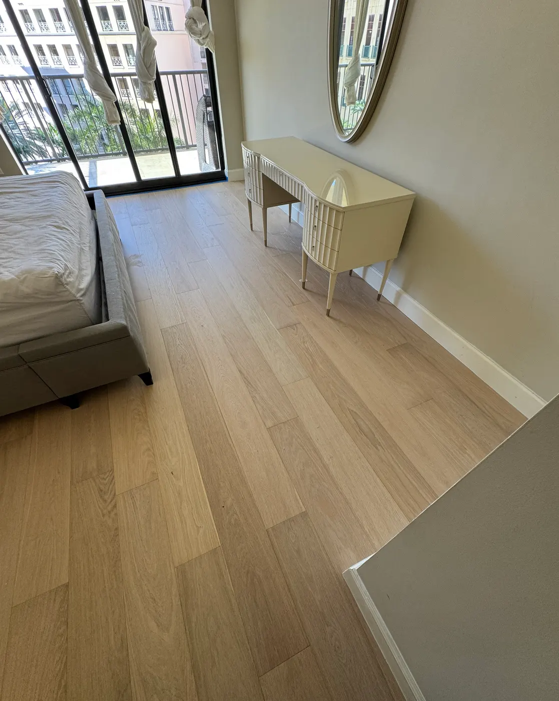 Bedroom with new light-toned wide-plank flooring and white baseboards next to a balcony door, installed by Colors Wood Floor