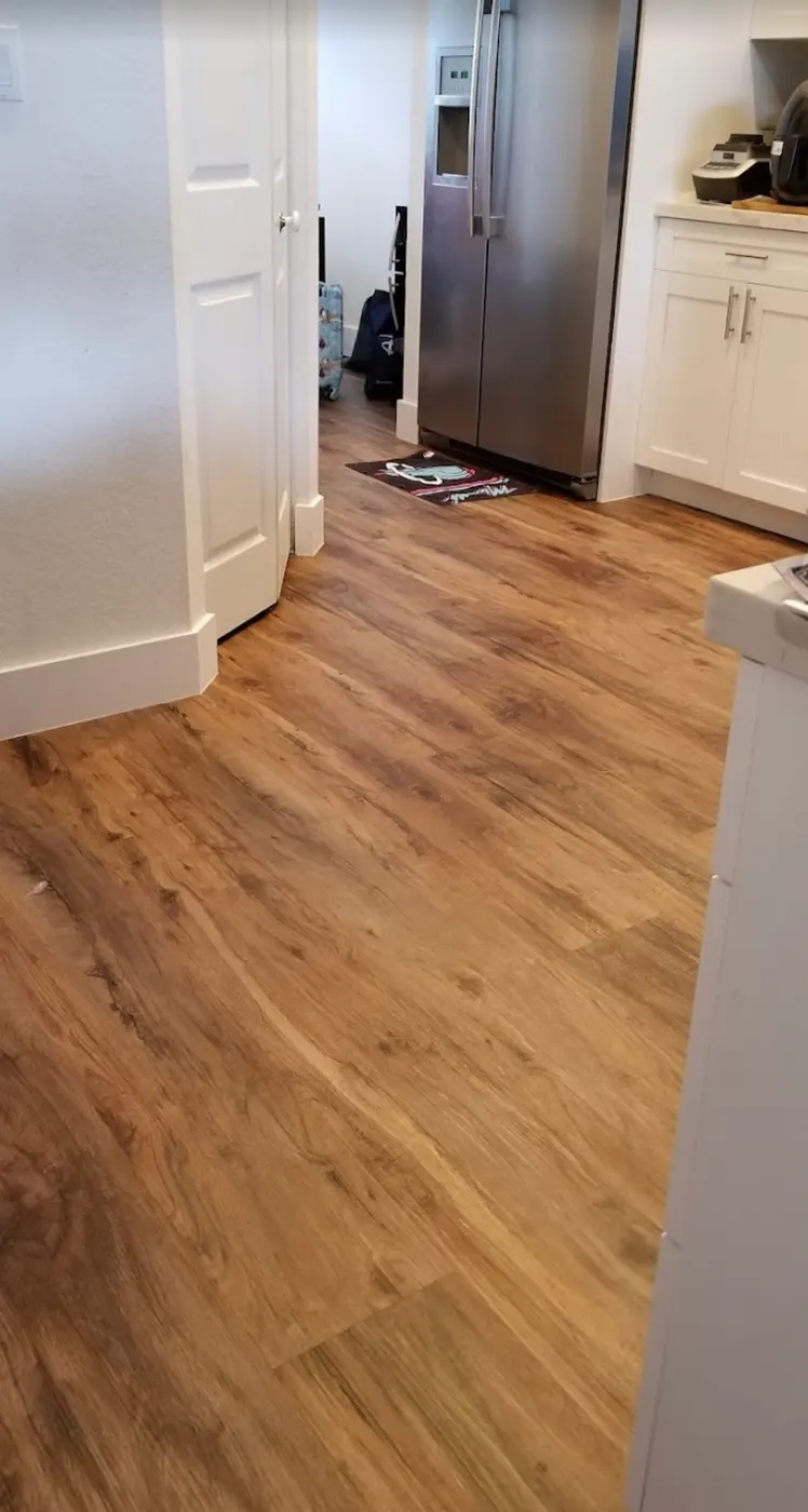 Wood-look vinyl plank flooring installed through a kitchen and hallway with white baseboards by Colors Wood Floor